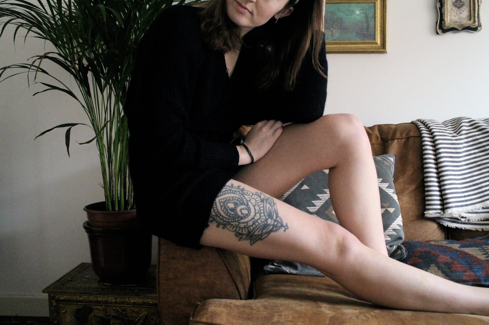 Girl in black dress with leg tattoo, sitting on edge of sofa