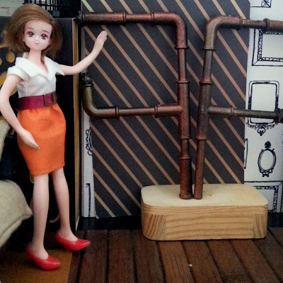 One-twelfth scale Takara compact doll in a modern miniature scene, pointing at two pipe shelving units set on top of a wooden pedestal.