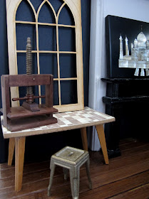Modern dolls' house miniature room corner with a vintage table, metal stool, book press and wooden window frame propped against the wall on the table.