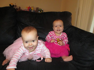 one year old twin baby girls, one in a light pink dress one in dark pink dress, sitting on black couch