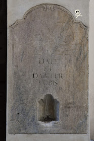 Antiguo limosnero de mármol, con grabado de calaveras y cita bíblica junto a la fecha de restauración del edificio.