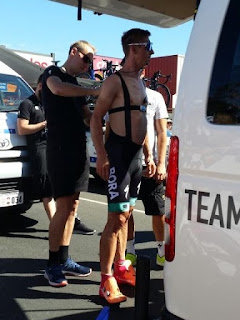 One of the Bora Hansgrohe riders is standing underneath the tailgate of the team bus with no jersey on.  One can see a heartrate monitor strapped around his chest and the braces for his shorts / knicks. A team technician is helping to hook up the radio pack. The rider is holding the earpiece in his mouth temporarily.
