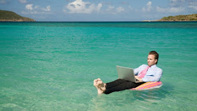 An office worker in a clear blue sea, floating on an inflatable chair, and working on a laptop.