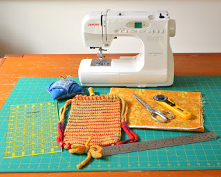 Sewing machine, materials and tools arranged on a cutting mat: quilter's rule, pin cushion with pins, a reel of sewing thread, a swatch of cotton quilting fabric, a rotary cutter, pair of scissors, steel rule and the WIP Project bag.