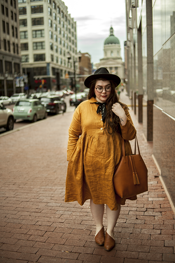 An outfit consisting of a black wide brim hat, an ochre linen baby doll dress, brown tote bag and brown d'oray flats.