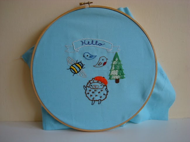 Embroidery of hedgehog wearing a beret, birds, hello on a banner, trees and bee