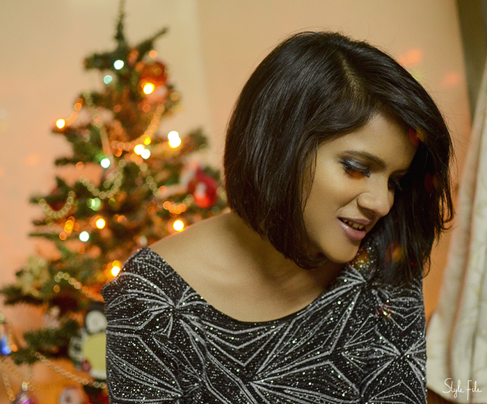 Image of a woman wearing silvery smokey eye makeup and fluttery lashes with a nude frost lipstick and graduated bob hairstyle on a silver glitter dress in front a Christmas tree