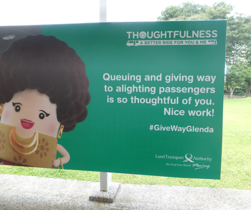 "Queuing and giving way to alighting passengers is so thoughtful of you. Nice work!" is displayed on a billboard. Below it is the hashtag "#GiveWayGlenda" and the words "Land Transport Authority: We Keep Your World Moving". Above it are the words "Thoughtfulness: A Better Ride for You & Me"