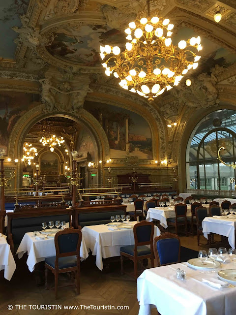 Ornate golden and blue interior design; tables are set with white linen and wine glasses. Elaborate paintings on the ceilings and in the vaults.