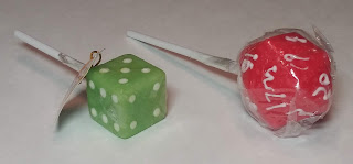 Two lollipops, both shaped like dice. The one on the left is a lime-green d6, and the one on the right is a red d20. They are both still in the cellophane.