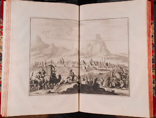 An illustration taking up a two-page spread, showing a caravan moving towards a distant city. 