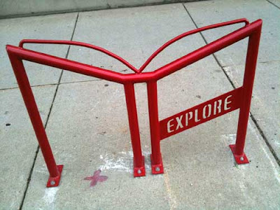 Red metal bike rack shaped like an open book with the word EXPLORE in open stencil letters