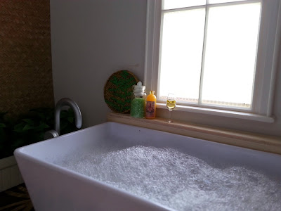 One-twelfth scale modern miniature bathroom with a floor-standing tub full of bubble bath, under a window, On the ledge is a tray with a fern pattern, a bottle of bath salts and one of bubble bath, plus a glass of sparkling wine.