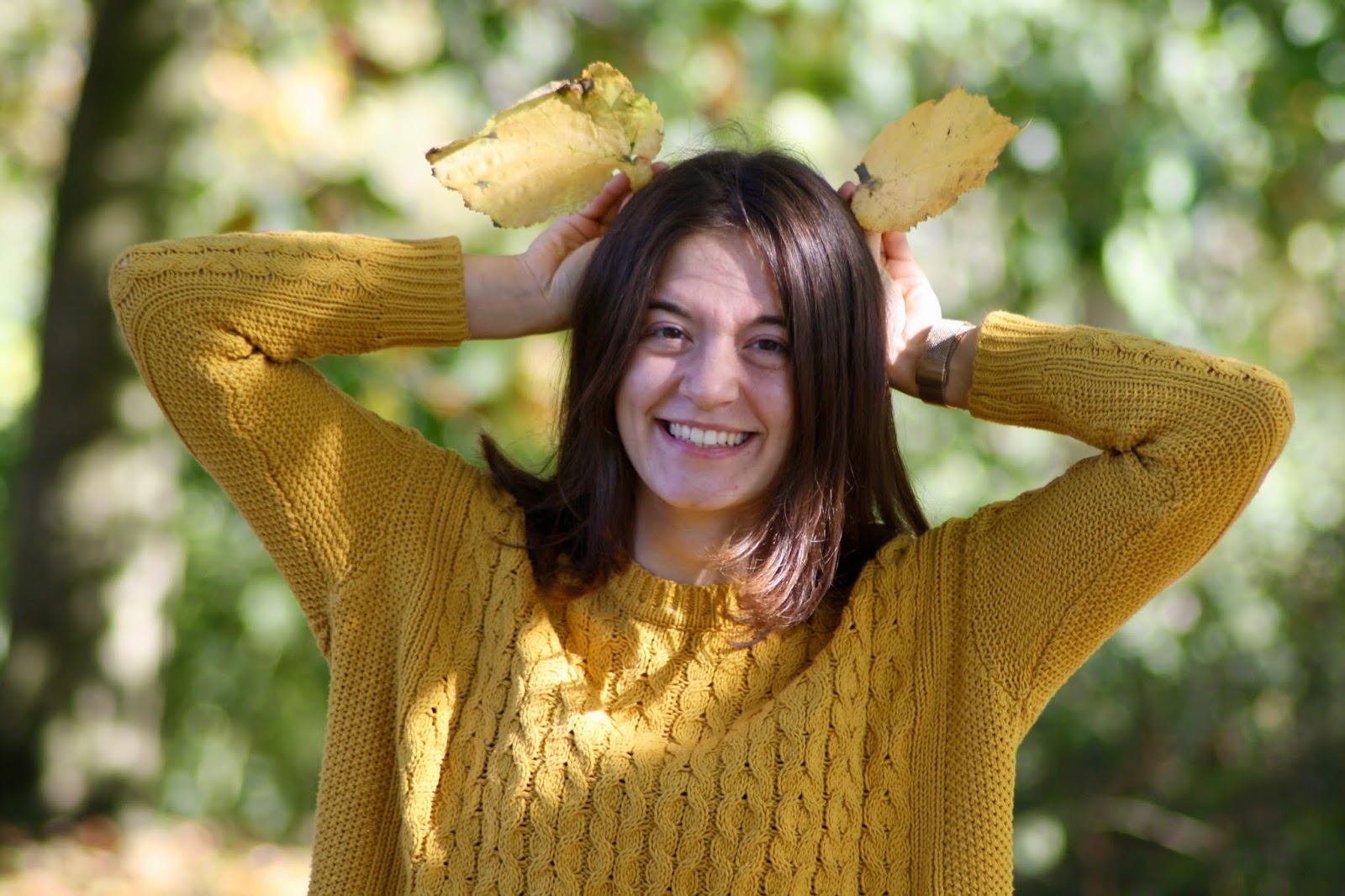abbey, a brunette blogger wearing a mustard knitted jumper, holds up two yellow autumn leaves on her head like ears