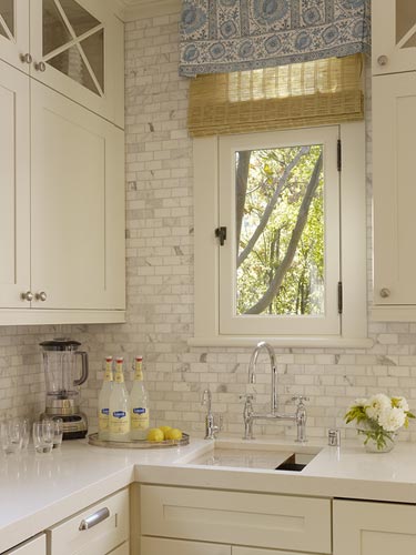 Close up of the Carrara marble backsplash in a kitchen with white cabinets, drawers and a small window with roman bamboo shades