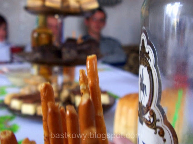 Rozmazane zdjęcie stołu imieninowego z widocznymi przekąskami, z naciskiem na charakterystyczne paluszki i fragment dekoracyjnej butelki, wywołujące uczucie domowego ciepła.
