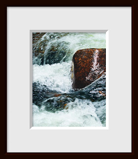 A framed photo of swirling and turbulent frigid mountain water careens by a large boulder