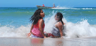 dos niñas sentadas a la orilla de la playa son golpeadas por una pequeña ola mientras una lancha pasa por detrás