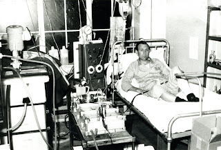 Man in pyjamas on bed with massively complex pumps and equipment all around him. He looks unconcerned by it. Blood reaches the machines from a shunt beneath bandages on his left leg. 