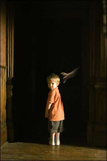Um garotinho loiro está parado em frente a um corredor escuro, de onde sai um braço pálido com aberta em direção à cabeça dele. O menininho está de costas, mas com o corpo virado para olhar para trás (onde está a câmera) e, por isso, não vê a mão ameaçadora saindo das sombras.