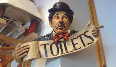 Carved Charlie Chaplin holding a role of toilet paper. Letters on the paper read Toilets