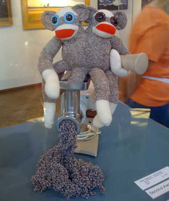 Two conjoined sock monkeys, one leg stuck in a meat grinder, brown and white yarn coming out the business end of it
