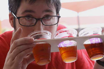 Young man with glasses drinking from a small plastic beer cup, which along with two other cups is embedded in a wooden paddle