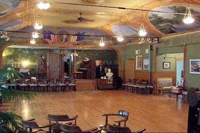 Green ballroom with wood paneling and painted scenes on the ceiling, a stage at one end of the room