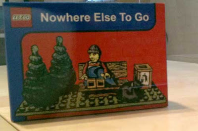 Solitary Lego bestubbled figure on a bench beside two pine trees, sack of belongings, trash can titled Nowhere Else to Go