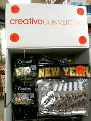 Photo of a white cardboard point-of-purchase display holding New Year's Eve decorations. The top of the display reads CreativeConverting