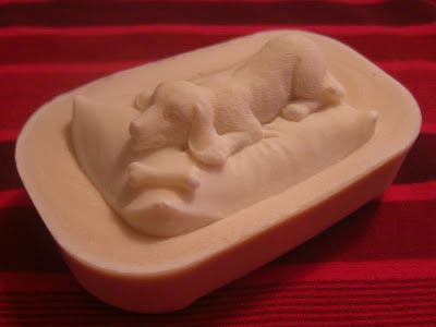 Picture of a bar of my goat milk soap with a puppy laying on a pillow beside a bone on top (the puppy is also soap!)