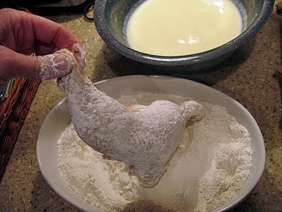 A photo of chicken being coated in the flour mixture with a bowl of egg mixture in the background.