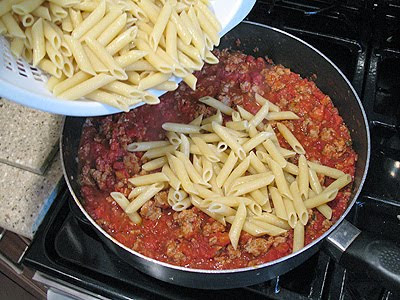 A photo of penne being poured over the sauce in a saute pan.
