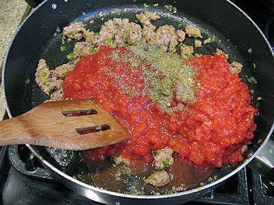 A close up photo of tomatoes, oregano, sage, and red pepper flakes added to the cooked sausage, shallots, and garlic mixture in a large saute pan.