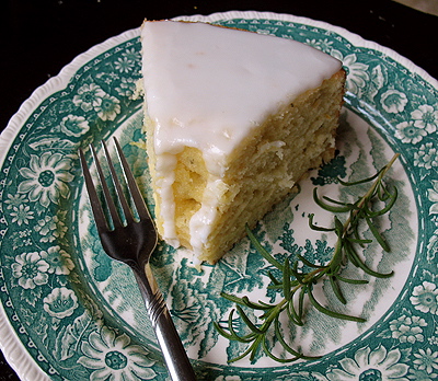 A close up photo of a slice of lemon rosemary olive oil cake resting on a decorative white and teal plate with a rosemary sprig on the side.